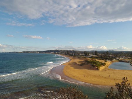 Panoramic view of the beach at Narrabeen, in Sydney, Australia, on a cloudy day with rough seas and orange sands and dunes leading to a small lagoon