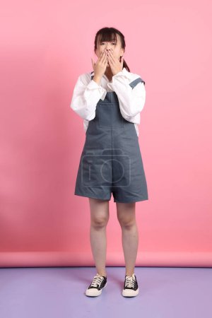 Cheerful lovely young asian woman in overalls casual clothes with gesture of Mouth close, Silent gesture isolated on pink background. St Valentine's Day, Women's Day, Birthday