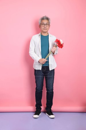 Homme asiatique âgé en tenue décontractée avec geste de tenir un bouquet de fleurs isolées sur fond rose. Saint Valentin, Anniversaire, Fête des femmes