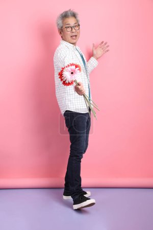 Asiatischer Senior in lässiger Kleidung mit Geste, einen Blumenstrauß auf rosa Hintergrund zu halten. Valentinstag, Frauentag, Geburtstag