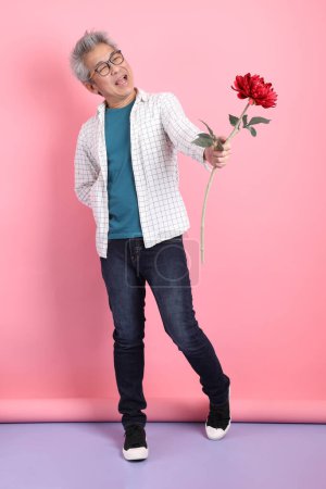 Asiatischer Senior in lässiger Kleidung mit Geste, einen Blumenstrauß auf rosa Hintergrund zu halten. Valentinstag, Frauentag, Geburtstag