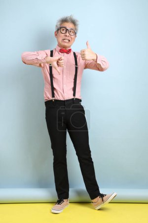 Asian senior man in black suspenders with red bow with gesture of thumbs down isolated on blue background. St Valentine's Day