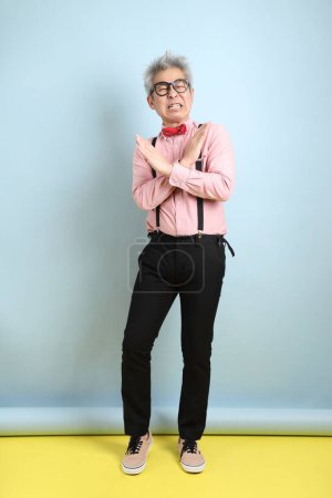 Asian senior man in black suspenders with red bow with gesture of stop, cross prohibition sign, forbid isolated on blue background. St Valentine's Day