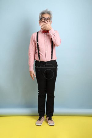 Asian senior man in black suspenders with red bow with gesture of Mouth close, Silent gesture isolated on blue background. St Valentine's Day