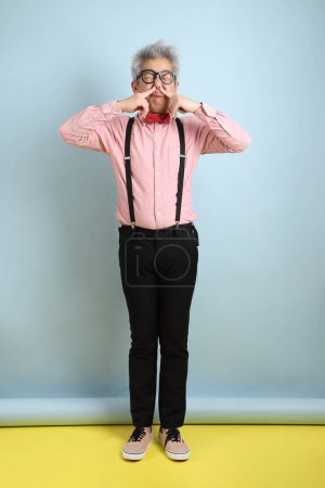 Asian senior man in black suspenders with red bow with gesture of smelling isolated on blue background. St Valentine's Day
