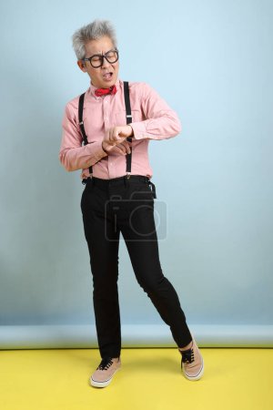 Asiatischer Senior in schwarzen Hosenträgern mit roter Schleife mit Geste des Wartens oder spät isoliert auf blauem Hintergrund. Valentinstag
