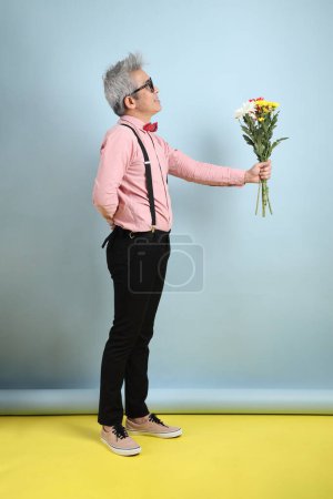 Asian senior man in black suspenders with red bow with gesture of holding a bouquet of flowers isolated on blue background. St Valentine's Day, Women's Day, Birthday