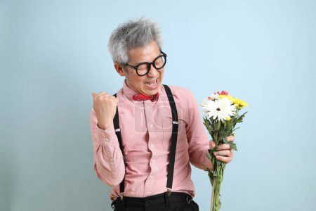 Asiatischer Senior in schwarzen Hosenträgern mit roter Schleife mit Geste, einen Blumenstrauß auf blauem Hintergrund zu halten. Valentinstag, Frauentag, Geburtstag