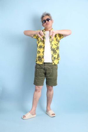HAPPY SONGKRAN DAY. Asian tourist senior man in summer clothing with gesture of thumbs down isolated on blue background. Songkran festival. Thai New Year's Day.