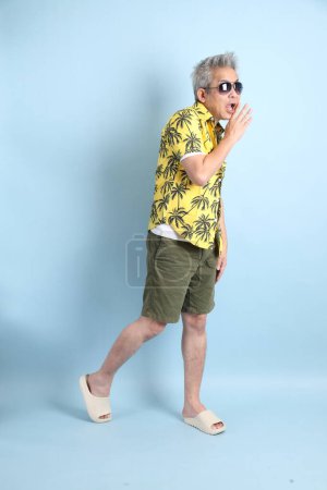 HAPPY SONGKRAN DAY. Asian tourist senior man in summer clothing with gesture of speak or whisper isolated on blue background. Thai New Year's Day.