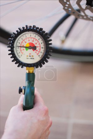 Mano sosteniendo un manómetro de neumático que indica 2,3 bar o 32 psi. Neumático borroso de bicicleta en el fondo. Contexto: inflar neumáticos de bicicleta, aire, monitoreo, seguridad de la bicicleta, servicio.