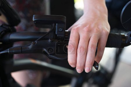 Vue rapprochée d'une main féminine saisissant un levier de frein de bicyclette, avec une commande E-bike visible sur le guidon.