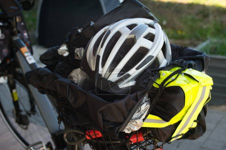 Rear view of an E-bike highlighting the bicycle luggage rack, which contains a bicycle helmet, reflective vest, bike lock, rain cover, and tarp. In the background, a bicycle tent is visible. The scene takes place outdoors within a fenced area.