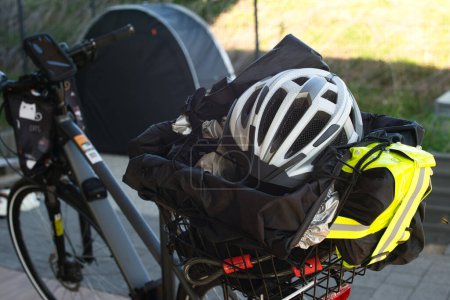 Rear view of an E-bike highlighting the bicycle luggage rack, which contains a bicycle helmet, reflective vest, bike lock, rain cover, and tarp. In the background, a bicycle tent is visible. The scene takes place outdoors within a fenced area.