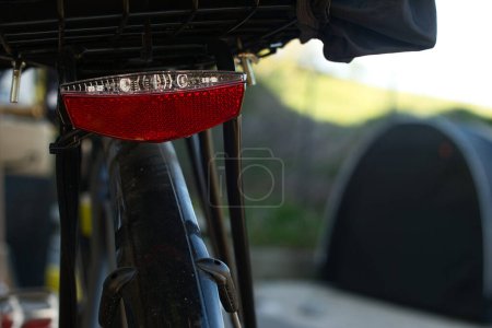 Close-up view of a red and white bicycle tail light mounted on a bike with a luggage rack. In the background, a black tent is blurred. The scene is outdoors.
