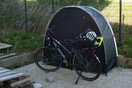 Electric bicycle with a luggage rack positioned in front of a bicycle tent. Various items such as a bicycle helmet, reflective vest, and rain cover are stored on the rack. The scene is outdoors within a fenced area with natural vegetation in the back