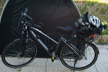 Vélo électrique avec porte-bagages placé devant une tente à vélos. Divers articles tels qu'un casque de vélo, un gilet réfléchissant et une housse de pluie sont stockés sur le support. La scène est à l'extérieur dans une zone clôturée avec de la végétation naturelle à l'arrière