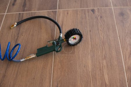Pressure gauge with a green handle, connected to a blue coiled hose, lying on wooden floor tiles.