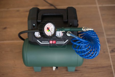 Green air compressor with pressure gauge connected to a compressor via a blue coiled hose, placed on wooden floor tiles.