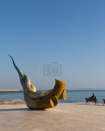 Durres, Albania - April 13th, 2024 - photo of a modern art sculpture by the sea, in the background a woman sitting at the bench by the sea, seagulls flying.