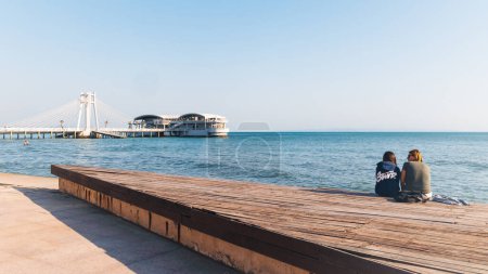 Foto de Durres, Albania - 13 de abril de 2024 - Foto del paseo marítimo de Durres con dos personas sentadas junto al mar sobre una plataforma de madera - Imagen libre de derechos