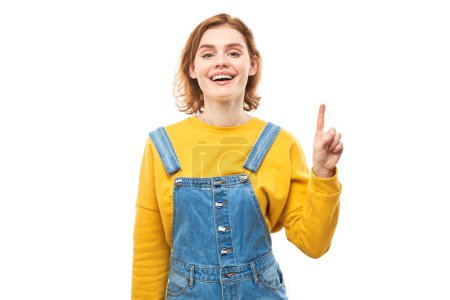 Foto de Retrato de una joven pelirroja positiva en jeans casuales sonriendo señalando con el dedo el espacio de copia vacío para texto o producto aislado sobre fondo blanco, banner publicitario - Imagen libre de derechos