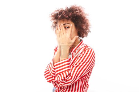 Foto de Mujer joven caucásica rizada espiando entre los dedos, cubriendo los ojos con palmas aisladas sobre fondo blanco. Concepto fobia - Imagen libre de derechos