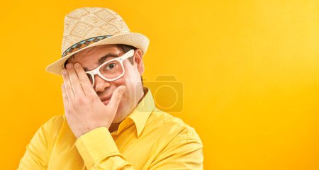 Téléchargez les photos : Homme au chapeau et lunettes regardant à travers les doigts, couvrant les yeux avec des paumes isolées sur fond jaune. Concept de phobie - en image libre de droit