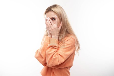 Téléchargez les photos : Jeune femme blonde regardant à travers les doigts, couvrant les yeux avec des paumes isolées sur fond blanc. Concept de phobie - en image libre de droit