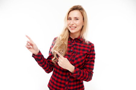 Photo for Joyful blonde girl in a plaid red shirt smiling points her finger at empty copy space for text or product isolated on white background, advertising banner - Royalty Free Image