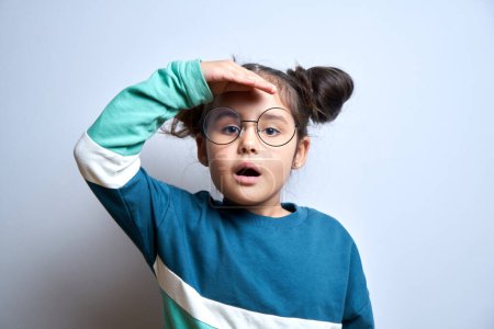 Photo for Little caucasian girl with glasses looks future, looking far away distance with hand over head isolated on white background. Search concept. - Royalty Free Image