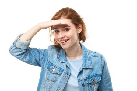 Photo for Redhead young woman in denim jacket looks future, looking far away distance with hand over head isolated on white background. Search concept. - Royalty Free Image