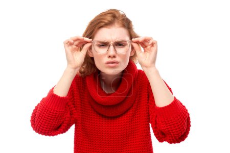 Foto de Mujer joven pelirroja con gafas entrecerrando los ojos mientras mira la cámara aislada sobre fondo blanco. Concepto problemas de visión - Imagen libre de derechos
