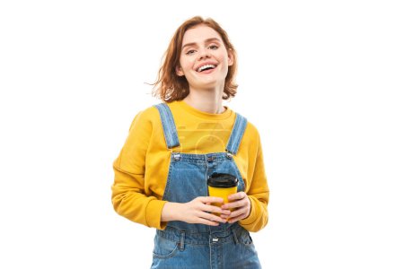 Foto de Enérgica pelirroja sosteniendo y bebiendo una taza amarilla de café para llevar, sonriendo y disfrutando de una bebida aislada sobre un fondo blanco - Imagen libre de derechos