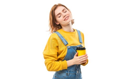 Foto de Enérgica pelirroja sosteniendo y bebiendo una taza amarilla de café para llevar, sonriendo y disfrutando de una bebida aislada sobre un fondo blanco - Imagen libre de derechos