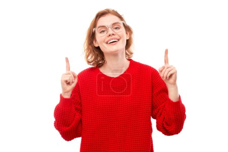 Téléchargez les photos : Portrait positive rousse jeune femme en pull de Noël rouge souriant pointant du doigt l'espace de copie vide pour le texte ou le produit isolé sur fond blanc, bannière publicitaire - en image libre de droit