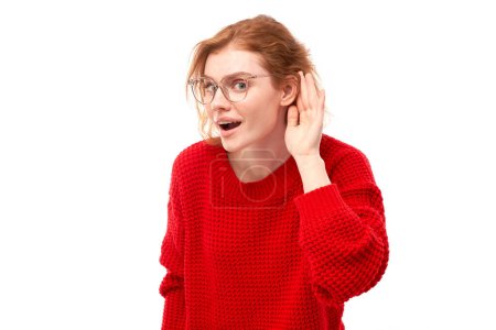 Téléchargez les photos : Jeune femme rousse en tenue décontractée souriant et tenant la main près de l'oreille, écoutant attentivement, entendant intensément isolé sur fond blanc - en image libre de droit
