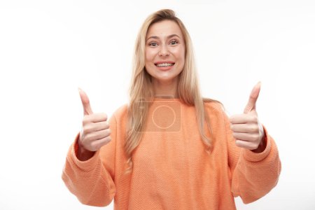 Téléchargez les photos : Heureuse fille blonde montre la main avec les pouces levés et sourit à la caméra isolée sur fond blanc - en image libre de droit