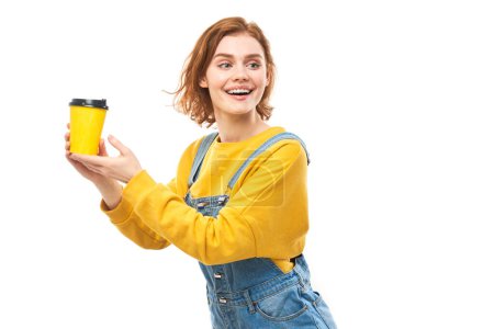 Téléchargez les photos : Fille rousse énergique tenant et buvant tasse jaune de café à emporter, souriant et savourant une boisson isolée sur fond blanc - en image libre de droit