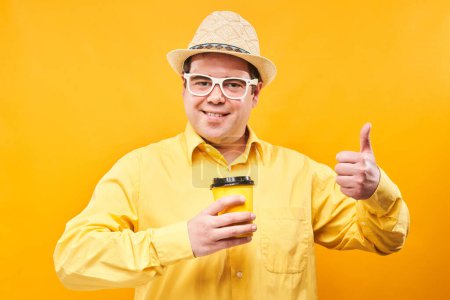 Téléchargez les photos : Homme drôle énergique tenant et buvant une tasse de café à emporter, souriant et savourant une boisson isolée sur fond jaune - en image libre de droit