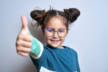 Téléchargez les photos : Petite fille caucasienne en lunettes montre la main avec les pouces levés et sourit à la caméra isolée sur fond blanc - en image libre de droit