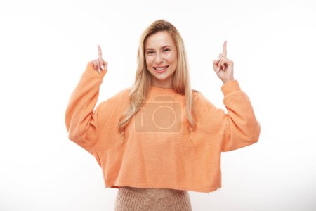 Téléchargez les photos : Joyeuse fille blonde en orange casual souriant pointe doigt à l'espace de copie vide pour le texte ou le produit isolé sur fond blanc, bannière publicitaire - en image libre de droit