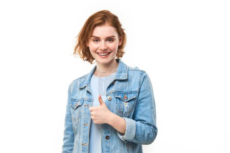Photo pour Jeune rousse femme montre la main avec les pouces vers le haut et sourit à la caméra isolée sur fond blanc - image libre de droit
