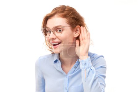 Téléchargez les photos : Jeune femme rousse en chemise d'affaires souriant et tenant la main près de l'oreille, écoutant attentivement, entendant intensément isolé sur fond blanc - en image libre de droit