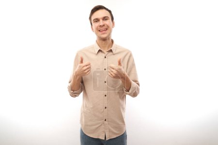 Téléchargez les photos : Jeune homme brune montre la main avec les pouces levés et sourit à la caméra isolée sur fond blanc - en image libre de droit