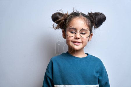 Téléchargez les photos : Portrait de petite fille intelligente rêveuse dans des lunettes et drôle de coiffure sourit et regarde loin isolé sur fond de studio blanc, heureux bel enfant - en image libre de droit