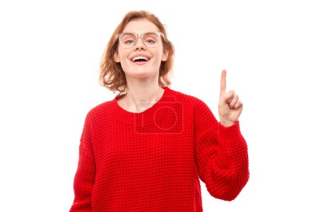 Téléchargez les photos : Portrait positive rousse jeune femme en pull de Noël rouge souriant pointant du doigt l'espace de copie vide pour le texte ou le produit isolé sur fond blanc, bannière publicitaire - en image libre de droit
