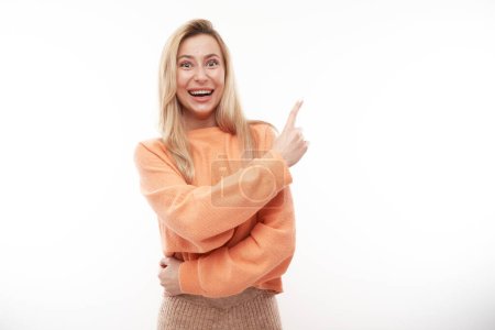 Téléchargez les photos : Joyeuse fille blonde en orange casual souriant pointe doigt à l'espace de copie vide pour le texte ou le produit isolé sur fond blanc, bannière publicitaire - en image libre de droit