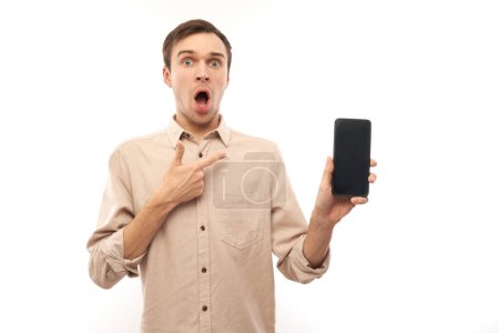 Foto de Retrato de un joven guapo hombre caucásico usando un teléfono móvil con la boca abierta y apuntando con el dedo a la pantalla en blanco aislado en el fondo del estudio blanco, expresión facial conmocionada - Imagen libre de derechos