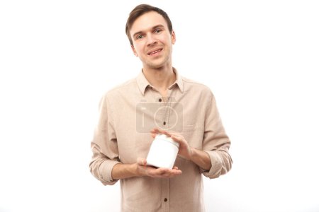 Foto de Retrato de un joven guapo sonriendo y sosteniendo un frasco blanco vacío con espacio de copia para texto en sus manos aislado sobre fondo blanco del estudio - Imagen libre de derechos